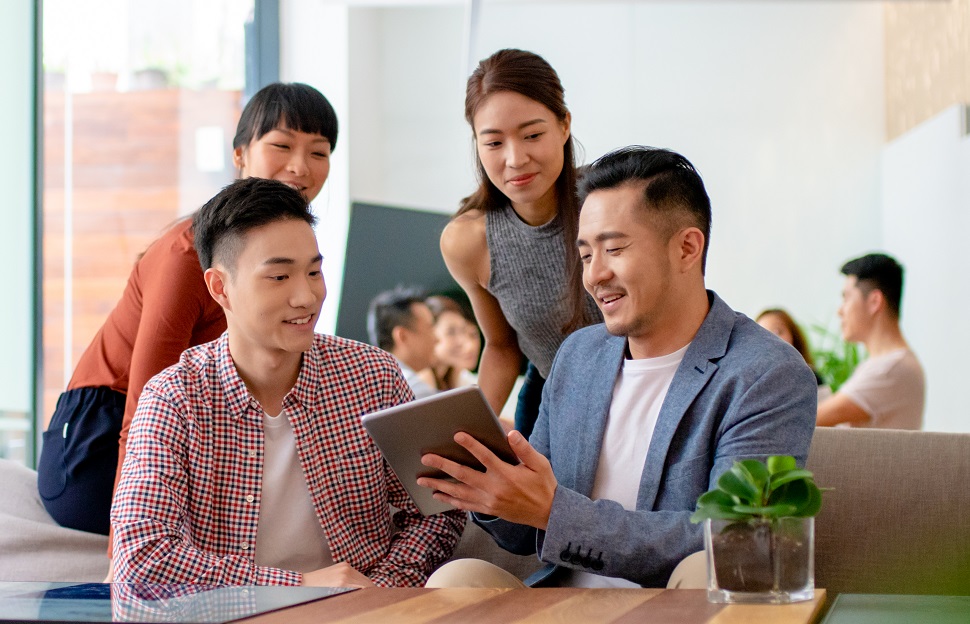 Two men and two women in smart casual clothing looking at one tablet, with another mixed gender group sitting in the background