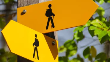 Two yellow hiking trail signs with arrows and backpack icons on a wooden post.