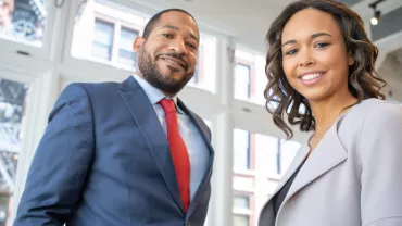 Two people smiling in a bright office with large windows.