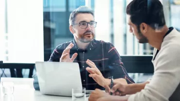 Two people having a discussion at a desk with a laptop and notepad.