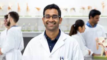 A person smiling in a lab coat with anatomical models in the background.