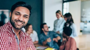 Smiling person in a checkered shirt stands in front of others in a modern office setting.