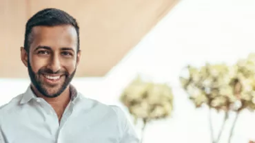 Person smiling in a white shirt, standing beside a glass wall with trees in the background.