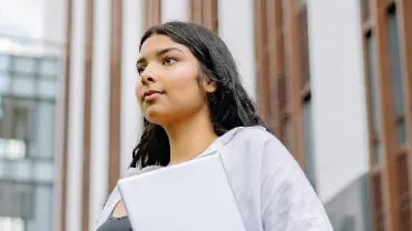 Person holding a laptop, standing in front of modern building.