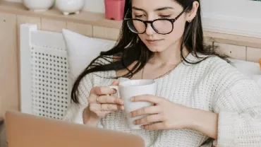 Person in a cozy room, reading on a laptop and holding a cup.