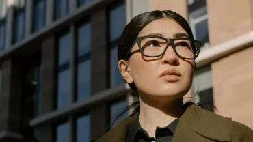 Female person with glasses stands in front of a modern building.