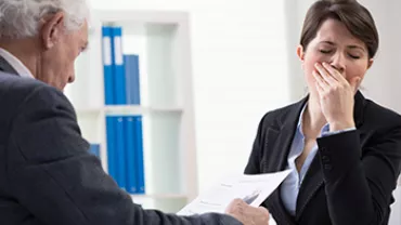 Person yawning while another hands them documents in an office.