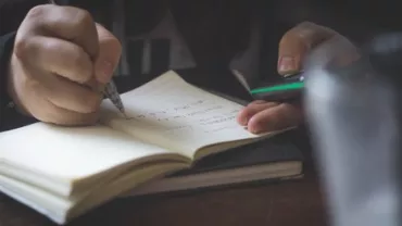 Person writing notes in a notebook while holding a phone.