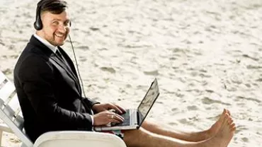 Person in a suit using a laptop on a beach chair, headphones on, smiling at the camera.