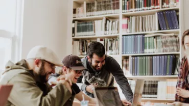 Group of people laughing and looking at a laptop in a library.