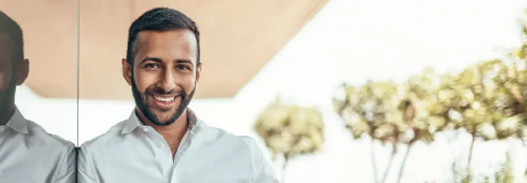 Person smiling in a white shirt, standing beside a glass wall with trees in the background.