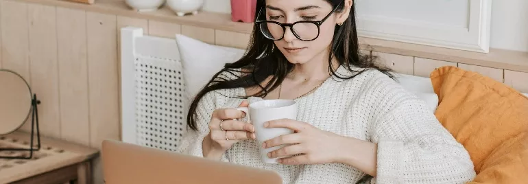 Person in a cozy room, reading on a laptop and holding a cup.