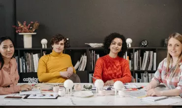 Four people sitting at a table in an office setting, surrounded by books and creative materials.