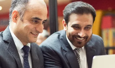 Two people in suits smiling while looking at a laptop.